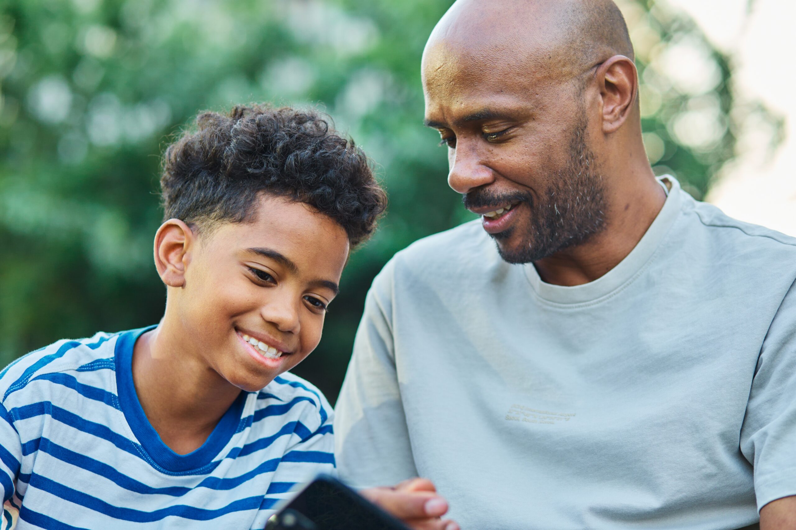 Father and son smiling together outdoors while looking at a smartphone, representing hope and connection through mental health treatments like TMS therapy in North Texas.